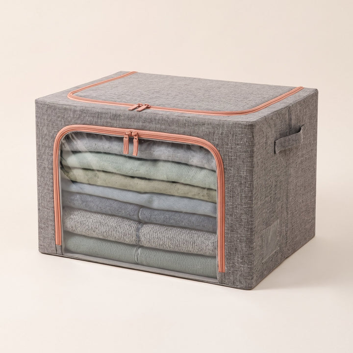 Caja guardarropa gris con ribetes naranjos, ventana frontal transparente y asas laterales, sobre fondo blanco hueso.