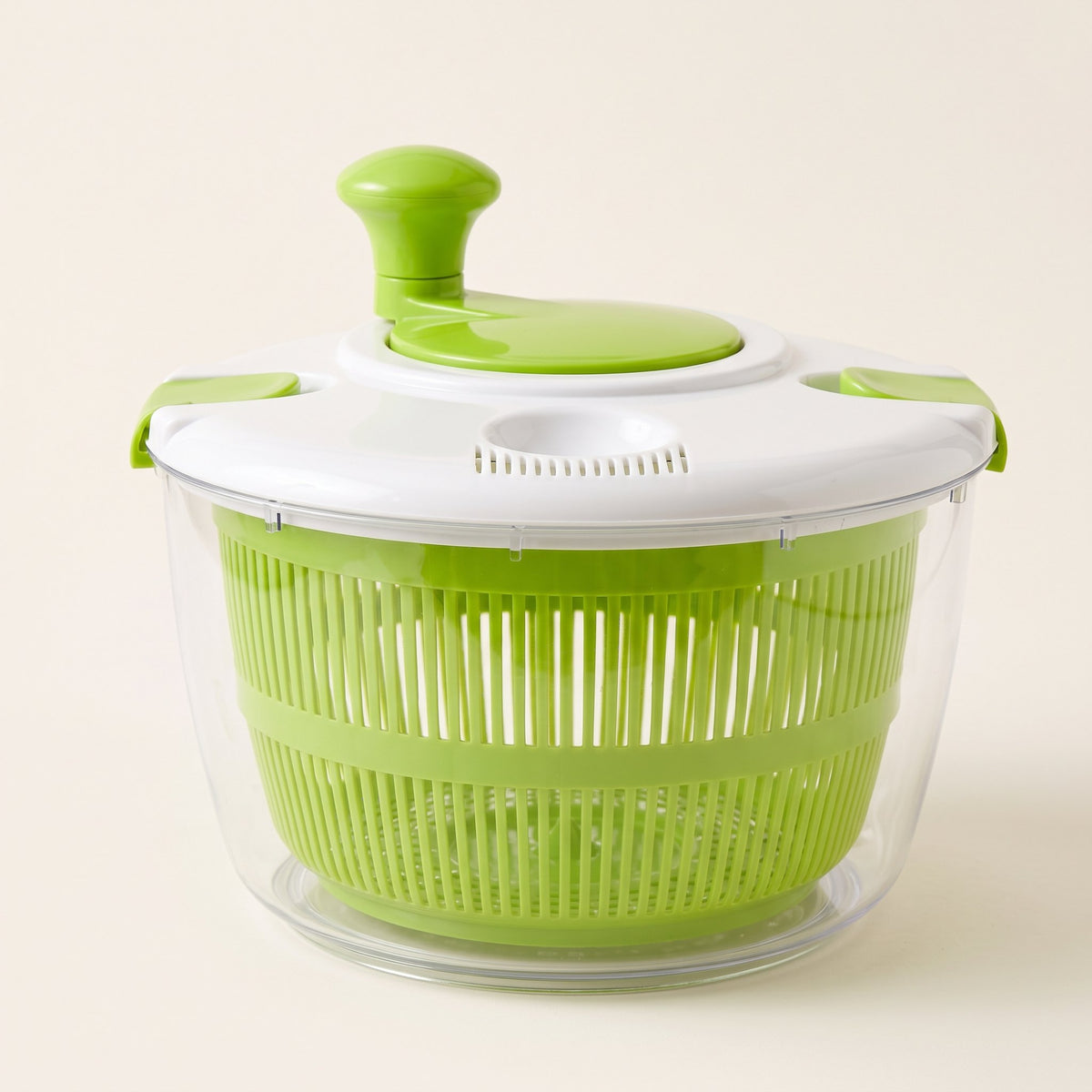 Centrifugadora de ensalada con bowl transparente, canasto verde y tapa blanca con manivela, sobre fondo blanco hueso.