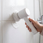 Mano limpiando el fragüe de los azulejos de la ducha con el cepillo eléctrico y espuma.