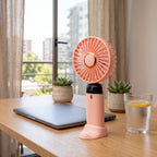 Ventilador portátil rosado en su base sobre un escritorio de madera junto a una ventana.
