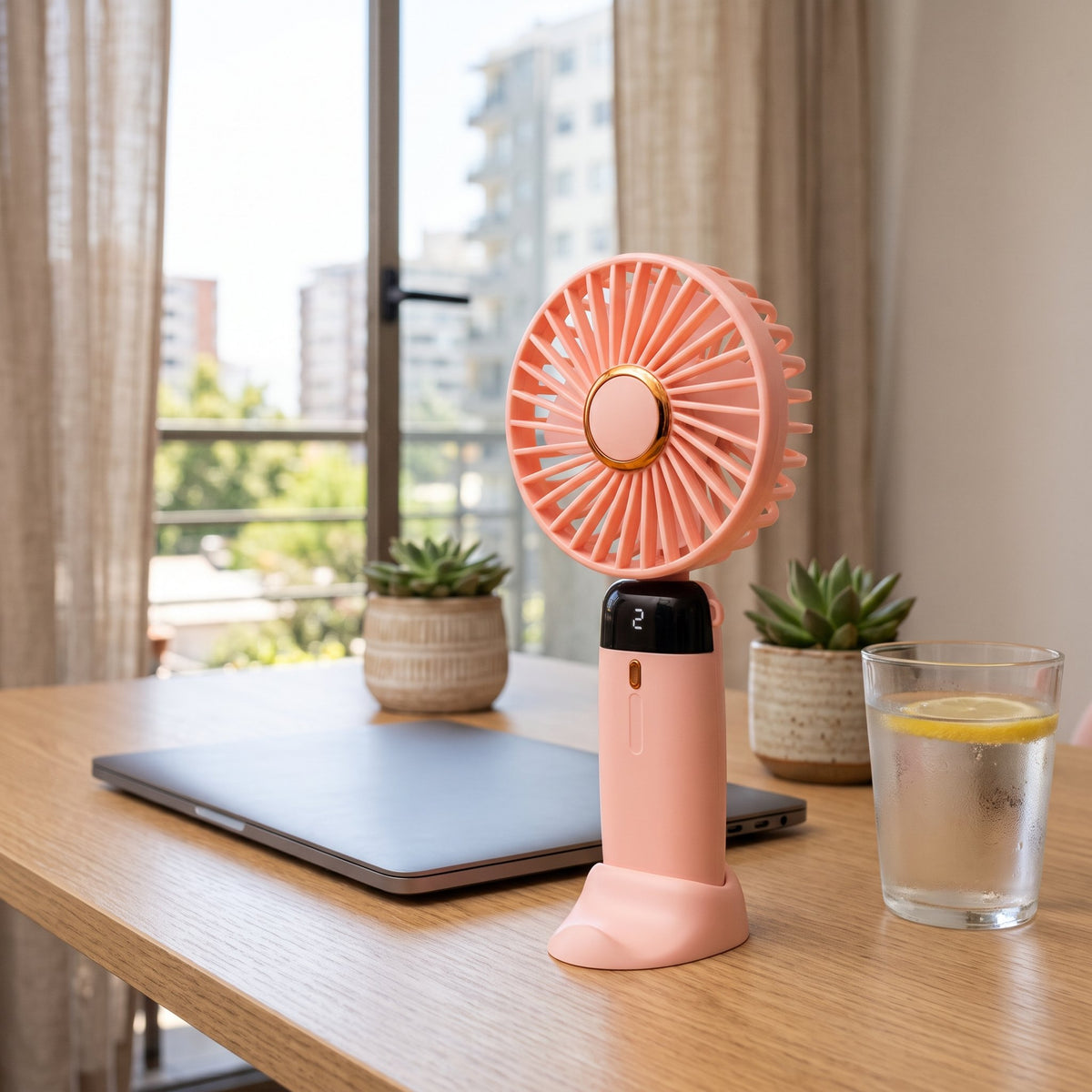 Ventilador portátil rosado en su base sobre un escritorio de madera junto a una ventana.