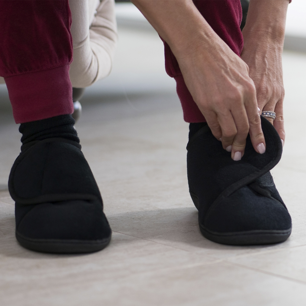 Mujer agachada ajustando la solapa de velcro de la pantufla negra que lleva puesta.